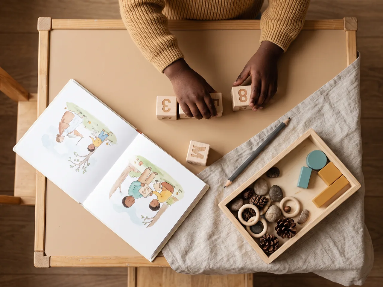 Overhead view of a small table with an open picture book, wooden number and letter blocks, a pencil, and a tray of natural materials.